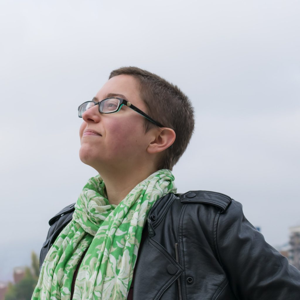 Image shows Fiction Editor Sam Corradetti outside, the sky seems overcast. They are wearing a linen scarf, a leather jacket and rectangular glasses. They are looking into the distance, their expression seems confident and satisfied.